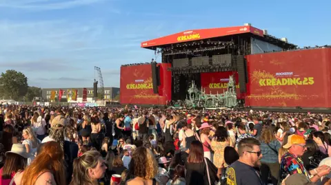 BBC An enormous crowd of people stand in front of a large stage. Massive red signs either side of the stage read 'Reading 25'. The sky is blue and largely cloudless and most of the people gathered are wearing summer clothes, with some wearing sunglasses and baseball caps.
