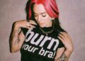 A woman with pink hair wearing a T-shirt that says burn your bra on it