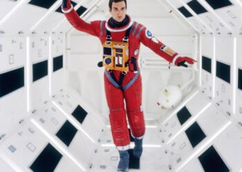 An astronaut in a red spacesuit walks inside a brightly lit, futuristic white corridor with geometric patterns. A white helmet floats nearby, suggesting a zero-gravity environment.