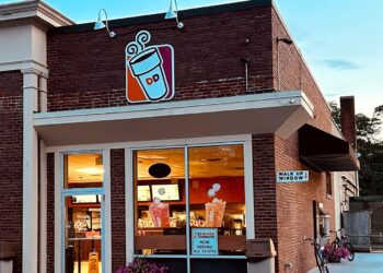 A Dunkin’ Donuts store with large front windows, a brick exterior, and a walk-up window sign. The inside lights are on, and there are pink flowers and a bicycle outside. The DD coffee cup sign is above the entrance.