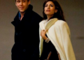 A man in a dark coat and a woman in a light coat and brown purse walk side by side, holding hands and looking in different directions. The background is plain and dimly lit.