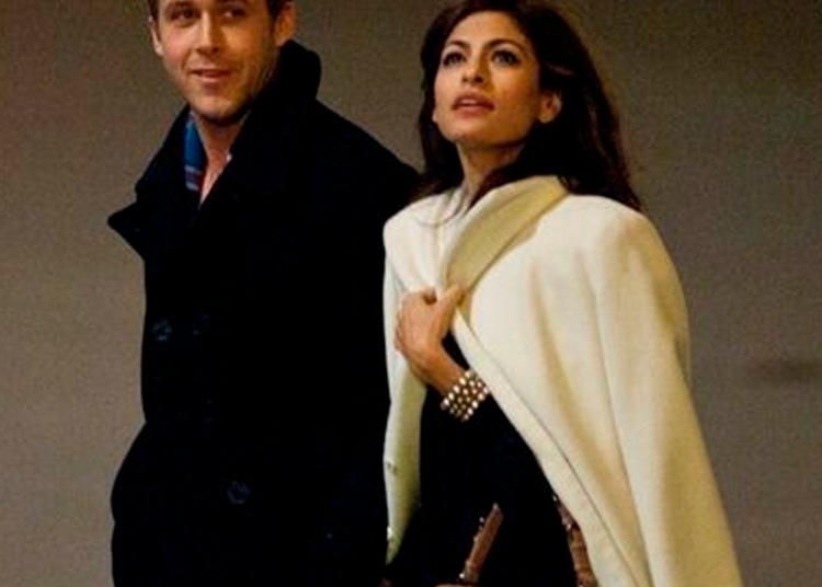 A man in a dark coat and a woman in a light coat and brown purse walk side by side, holding hands and looking in different directions. The background is plain and dimly lit.