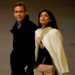 A man in a dark coat and a woman in a light coat and brown purse walk side by side, holding hands and looking in different directions. The background is plain and dimly lit.