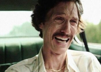 A man with tousled hair and a mustache sits in a car, wearing a white shirt and gold necklace, displaying an intense, emotional expression with teary eyes and an open-mouthed grimace.