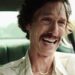 A man with tousled hair and a mustache sits in a car, wearing a white shirt and gold necklace, displaying an intense, emotional expression with teary eyes and an open-mouthed grimace.