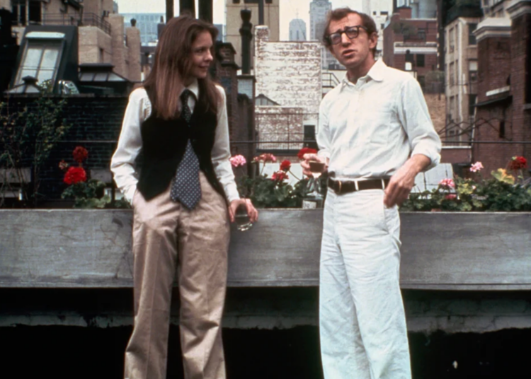 A woman in a vest, tie, and wide-legged pants stands beside a man in a white shirt and pants. They are outdoors on a city rooftop with flowers and buildings in the background, both holding drinks and talking.