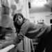 A woman in a sweater washes dishes at a kitchen sink, glancing over her shoulder and smiling. The kitchen is cluttered, and a bright ceiling light illuminates the scene.