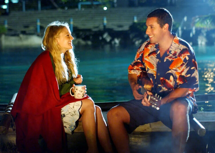 Man playing guitar for a woman wrapped in a red blanket as they sit by the water at night in 50 First Dates.