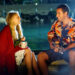 Man playing guitar for a woman wrapped in a red blanket as they sit by the water at night in 50 First Dates.