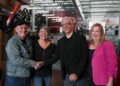 A portrait of two men and two women standing in warehouse with technical equipment behind them as the two men shake hands.