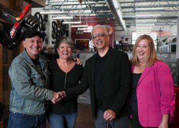 A portrait of two men and two women standing in warehouse with technical equipment behind them as the two men shake hands.