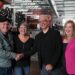 A portrait of two men and two women standing in warehouse with technical equipment behind them as the two men shake hands.