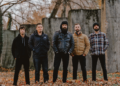 Members of August Burns Red standing outdoors in a fall setting, posing together in front of weathered concrete walls with fallen leaves on the ground.