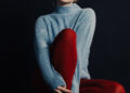 Actress Anna Baryshnikov photographed by Daniela Spector. She is wearing a baby blue mock neck sweater with red opaque tights. She is sitting on a stool against a black background.