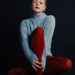Actress Anna Baryshnikov photographed by Daniela Spector. She is wearing a baby blue mock neck sweater with red opaque tights. She is sitting on a stool against a black background.