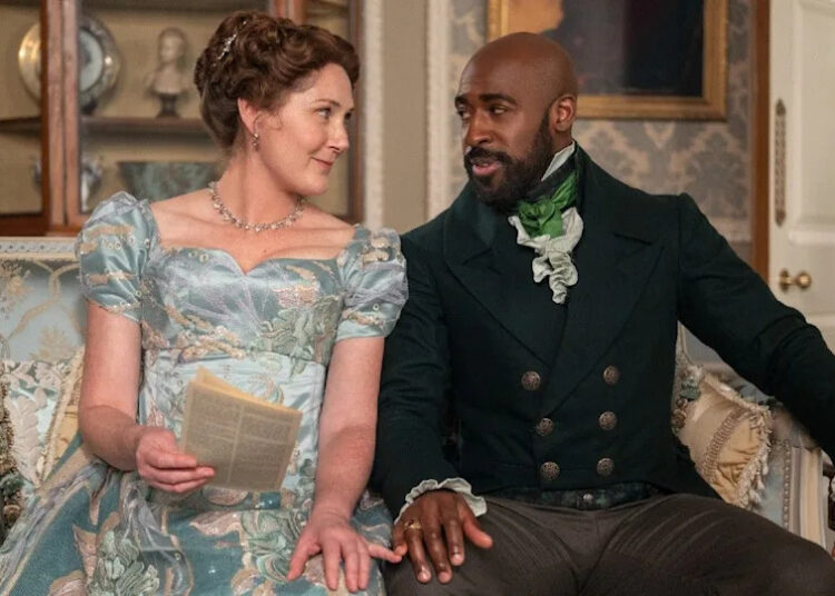From left to right: Ruth Gemmell as Lady Violet Bridgerton and Daniel Francis as Lord Anderson. They're sitting together on a couch and touching pinkies. .