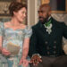 From left to right: Ruth Gemmell as Lady Violet Bridgerton and Daniel Francis as Lord Anderson. They're sitting together on a couch and touching pinkies. .