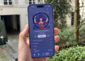 A hand holding an iPhone 17 Pro Max. The phone shows the new iOS 26.4 Apple Music design for the album's list.