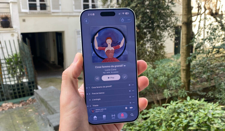 A hand holding an iPhone 17 Pro Max. The phone shows the new iOS 26.4 Apple Music design for the album's list.