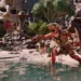 Screenshot of an enemy camp in Avowed in a desert area with multiple monsters. A lizard creature strikes out with a polearm as it roars.