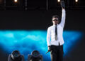 Baby Keem on stage wearing a white shirt, black tie, and gloves, raising one hand. Background features blue lighting.