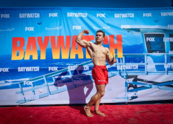 A muscular man flexing on a red carpet with a banner that says Baywatch across it.