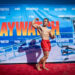 A muscular man flexing on a red carpet with a banner that says Baywatch across it.