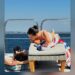 Two figures relaxing on a sunbed by the water.