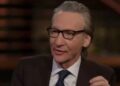 An individual dressed in formal attire, consisting of a suit and tie, is seated and appears to be engaged in conversation. The p
