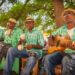 Four musicians in straw hats and matching green plaid shirts play instruments under a tree. One plays maracas, one a guitar, and the other two use percussion instruments.