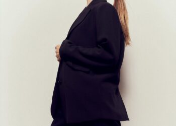a figure in a tailored black outfit stands side profile against a light background