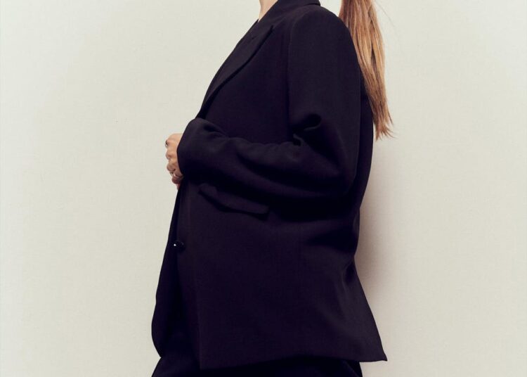 a figure in a tailored black outfit stands side profile against a light background