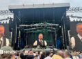 A picture taken from the crowd of three screens showing Bruce Springsteen playing on stage. Springsteen has short grey hair and is singing into a mic, he is wearing a white shirt, black tie and waistcoat.