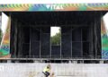 Image of the Vital stage set up at Boucher Road Playing Fields, a man working is riding his bike passed the stage. He has a hi vis jacket on. The stage is metal and has a sign reading VITAL on the top of the stage.