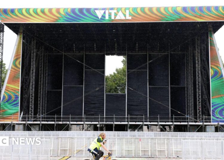Image of the Vital stage set up at Boucher Road Playing Fields, a man working is riding his bike passed the stage. He has a hi vis jacket on. The stage is metal and has a sign reading VITAL on the top of the stage.