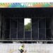 Image of the Vital stage set up at Boucher Road Playing Fields, a man working is riding his bike passed the stage. He has a hi vis jacket on. The stage is metal and has a sign reading VITAL on the top of the stage.