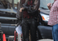 Will Ferrell in costume as a character with long blonde hair and a beard, wearing a black sheer shirt, a red and black vest, and black leather pants, on a film set.