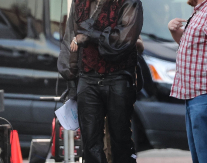 Will Ferrell in costume as a character with long blonde hair and a beard, wearing a black sheer shirt, a red and black vest, and black leather pants, on a film set.