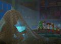 In a scene from Disney and Pixar’s Toy Story 5, we are looking at a darkened bedroom from the head of a twin bed. Directly in front of us, on the left side of the image, a mysterious person, probably a child, is silhouetted beneath a blue polka-dot sheet, holding a computer tablet that has them entranced. At the end of the bed, peeking over the foot of the bed and lit by a beam of moonlight, are many of the Toy Story characters, including Woody, Jessie, and Buzz.