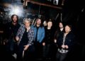 2026 band photo of six members of Foo Fighters standing together with guitars and keyboards in a studio setting, posing confidently as a group.