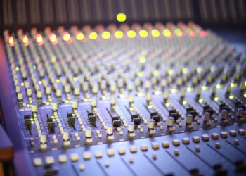 A professional music mixing console with numerous knobs and glowing indicator lights in a recording studio.