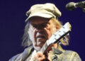 Neil Young is playing a guitar on stage while wearing a green cap and checkered jacket. He has long hair and is standing next to a microphone.