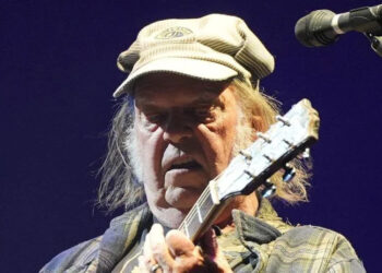 Neil Young is playing a guitar on stage while wearing a green cap and checkered jacket. He has long hair and is standing next to a microphone.