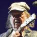 Neil Young is playing a guitar on stage while wearing a green cap and checkered jacket. He has long hair and is standing next to a microphone.