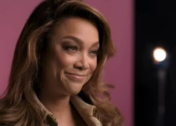 Person smiling softly, wearing a button-up top, in a studio setting with blurred lights in the background