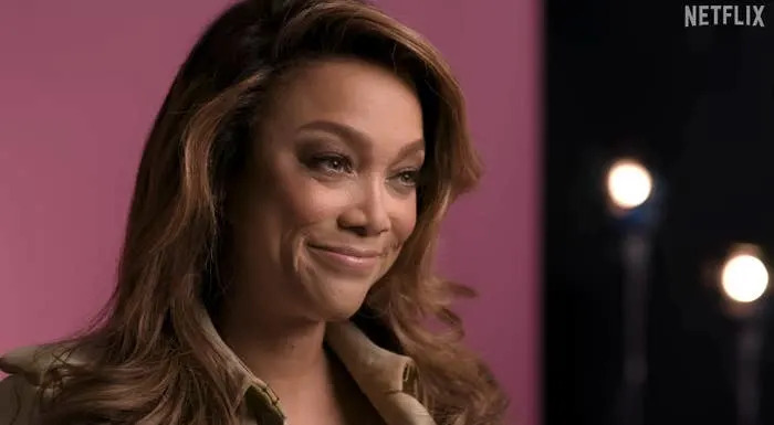 Person smiling softly, wearing a button-up top, in a studio setting with blurred lights in the background