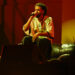 J. Cole performing on stage, wearing a white jersey, sitting on a couch, holding a microphone under red stage lighting.