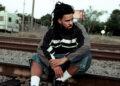 J. Cole sitting on train tracks, wearing a striped jacket and denim shorts, with a pensive expression.