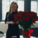 A blonde woman in a black dress reads the card from a giant bouquet of red roses in a vase on her desk.