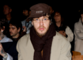 Jack Harlow wearing glasses, a brown hat, and a scarf sits in a crowd. Others around him have various styles, including tattoos and sunglasses.